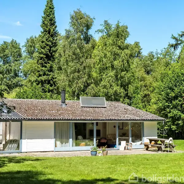 Et hvidt hus med fladt tag står stille på en græsplæne omgivet af træer. En terrasse foran huset har havemøbler og blomsterkrukker.
