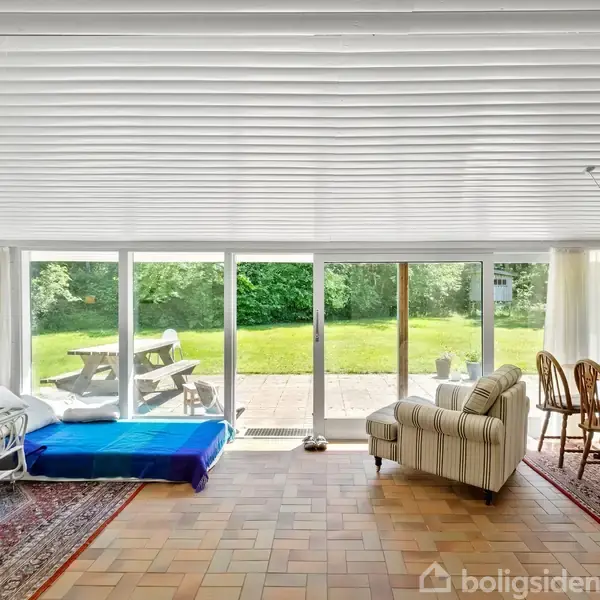 Stue med sofaer og spisebord, handlingen er afslapning og spisning, konteksten er et lyst rum med panoramavinduer, der åbner ud til en græsplæne og et picnicbord udenfor.