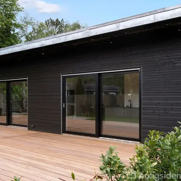 Sort træhus med store glasskydedøre står på en rummelig træterrasse. Baggrunden består af grønne træer og buske, hvilket skaber en naturlig og rolig atmosfære.
