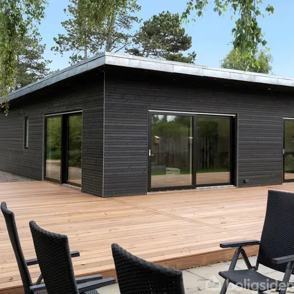 Et moderne, sort træhus med store glaspartier står på en rummelig træterrasse. Tre sorte stole er placeret på terrassens flisebelagte område. Omgivet af frodig grøn natur.