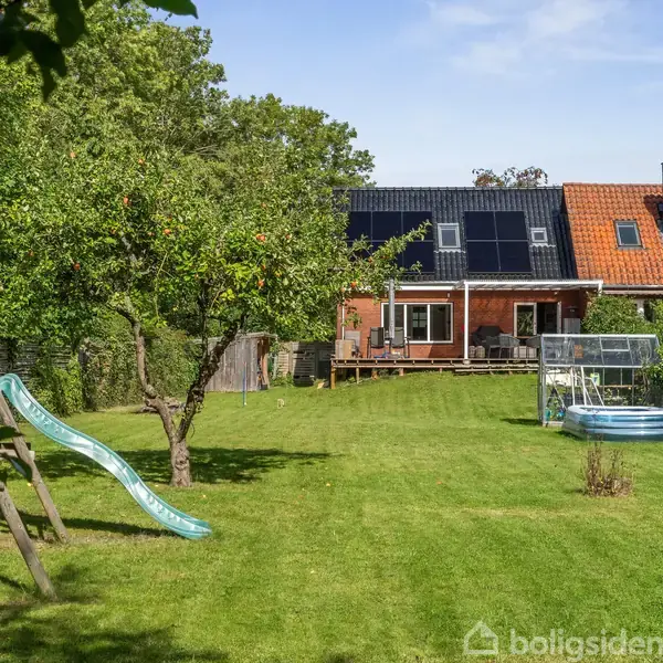 Hus med solpaneler på taget, store vinduer, og en terrasse i en stor have. Der er en barneslæde og rutsjebane til venstre samt et oppusteligt bassin til højre.