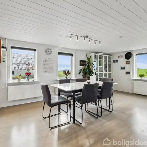 Spisebord med seks stole står i et lyst køkken-alrum. Store vinduer med udsigt til grønne områder og dekoreret med blomster. Vægge har ure og dekorative skilte.