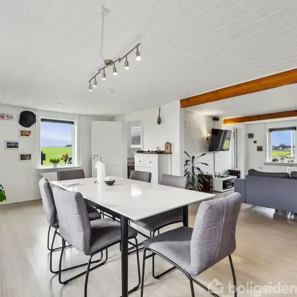 Spisebord med stole placeret i et lyst, moderne køkken-alrum. Store vinduer giver udsigt til grønne marker, og der er både sofaområde og forskellige dekorationer på væggene.