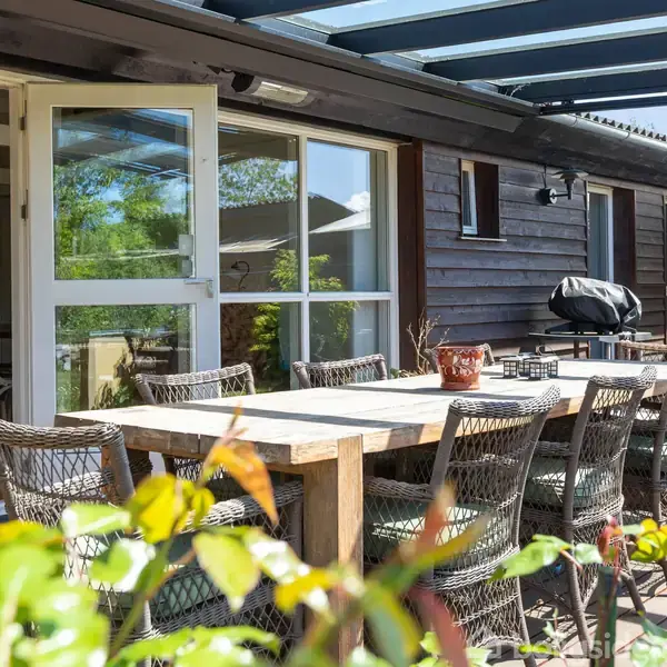 Spisebord med kurvestole står på træterrasse. Bordet er dækket af en glasoverdækning. Står foran moderne træhus med store vinduer og åben dør til køkkenområde.