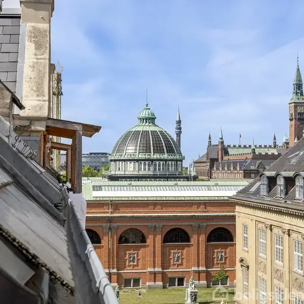 Bygninger med skrå tage omkranser en gård, mens en stor, kuppelformet struktur dominerer baggrunden under en klar, blå himmel. Gamle mursten og moderne arkitektur blandes.