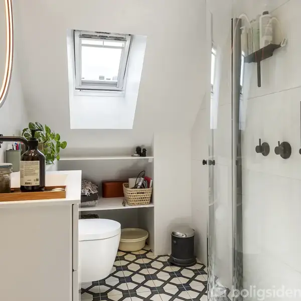 Et moderne badeværelse indeholder en håndvask med spejl, et væghængt toilet og en brusekabine. Der er planter og hylder med kurve. Det geometriske gulvfliser skaber et elegant udseende.