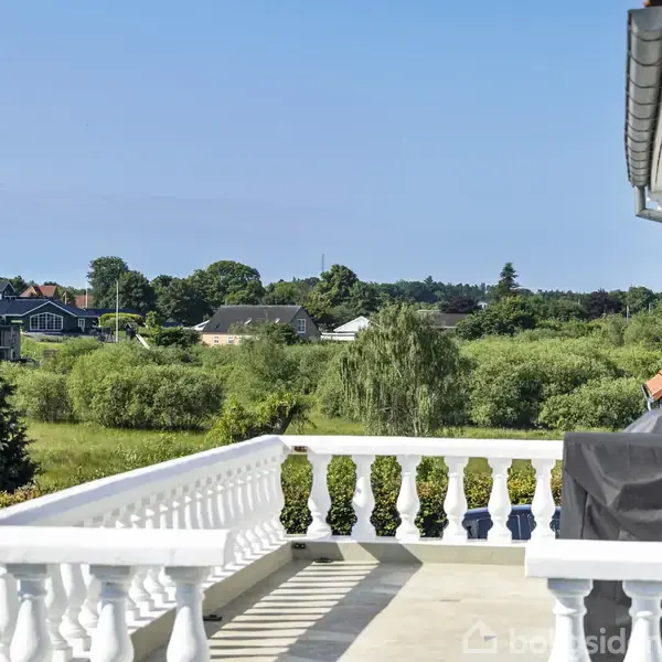 Hvid balkon med gelænder, stående stille, med udsigt over en grøn landskabsudsigt med træer og huse i baggrunden, under en klar blå himmel.
