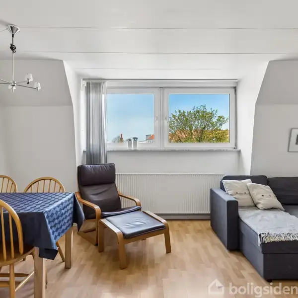 Stue med en grå hjørnesofa til højre, et spisebord med fire træstole til venstre. Et stort vindue i midten med udsigt til et træ udenfor.