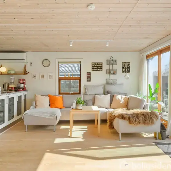 Lys stue med hvid sofa og orange pyntepude, placeret ved store glasdøre, som fører ud til en terrasse. Trægulv og hvidt køkken til venstre. Ingen tekst til stede.