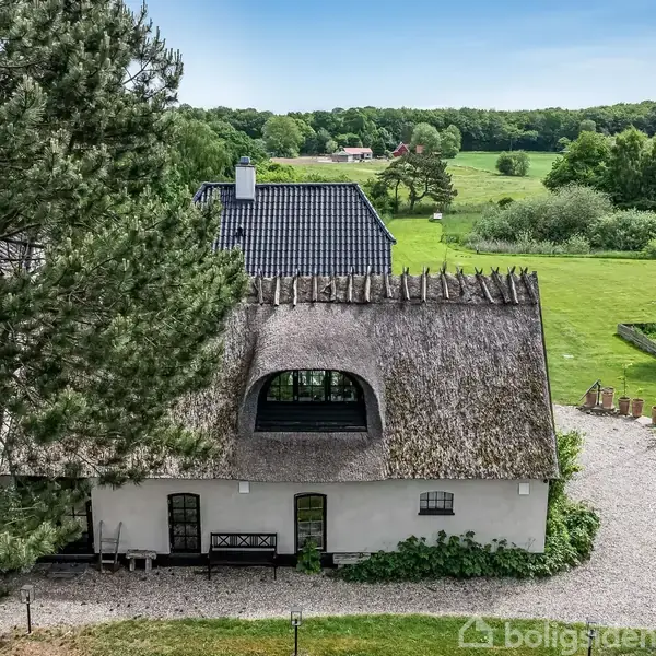 En stråtækt bygning står stille i en frodig, grøn have med grusgang og flagstang, omgivet af træer og en stor plæne, med skov i baggrunden.