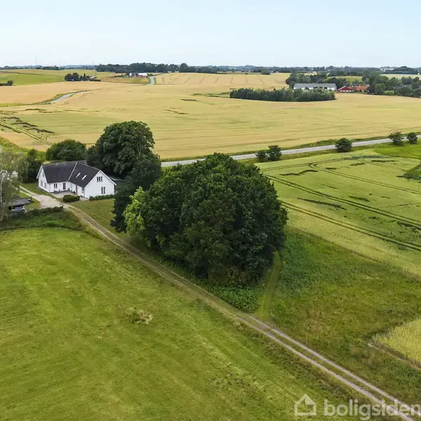Et hvidt hus ligger på landet, omgivet af grønne marker og træer. En vej løber forbi huset, og i baggrunden ses åbne marker og skovområder.