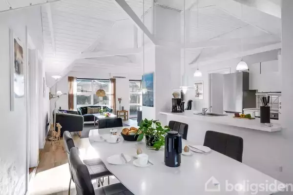 Spisebord dækket med kaffekande og kopper, strækker sig gennem en lys, moderne stue med sofaer. Store vinduer slipper dagslys ind i rummet, som også har en åben køkkenområde.