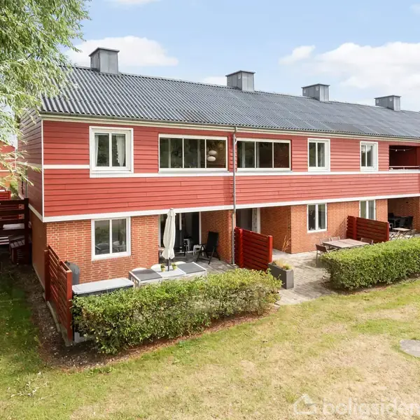 Rækkehuse i røde mursten med hvid ramme; terrasseområder foran adskilt af hække. Plæne og træer danner en grøn, åben baggrund.