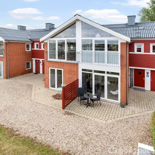 Rødt murstenshus med store vinduespartier står stille i et gårdmiljø. Grusbelægning omgiver huset med en terrasse foran. Huset har en moderne arkitektur med skrånende tag og altaner.
