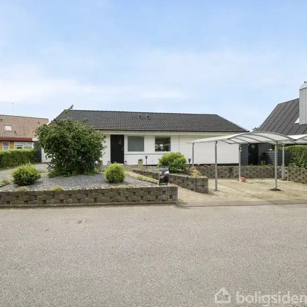Hus med fladt tag står stille. Forhave med indgang og overdækket carport. Nabohuse på begge sider, beliggende i et villakvarter. Grå belægningssten dekorerer indkørslen.