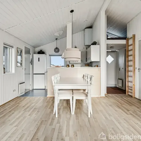 En hvid spisebordssæt står centralt i et lyst køkken-alrum med trægulv og hvide vægge. I baggrunden ses køkkenelementer, et køleskab og en emhætte i et moderne skandinavisk design.