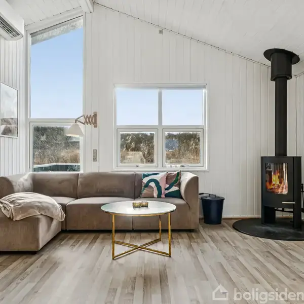 Brændeovn i hjørnet af en stue med lys trægulv. Ved siden af er der en beige sofa med puder, foran et rundt sofabord, og store vinduer.