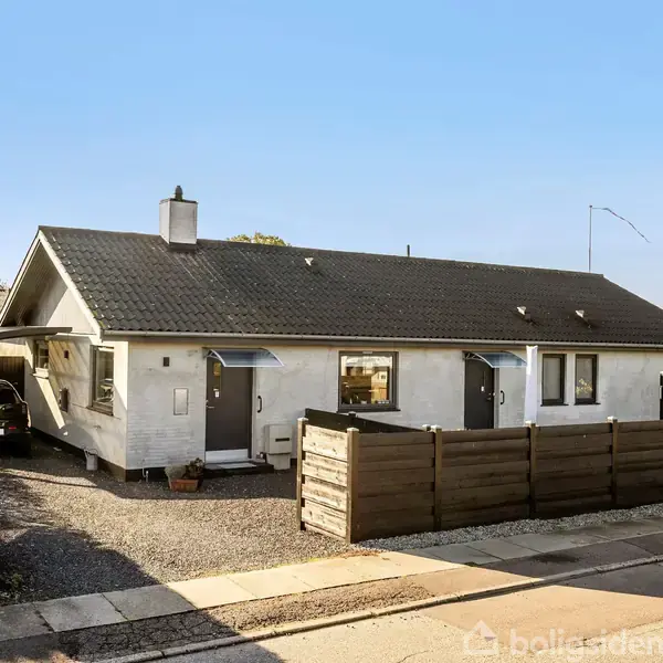 Et enkelt hvidt enfamiliehus med mørkt tag står ved siden af en carport med en bil. En hæk og et træhegn omgiver grunden i et villakvarter.