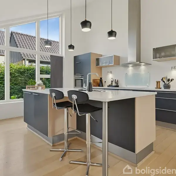 En moderne køkkenø med to barstole står centralt, omgivet af hvide skabe og stålapparater. Store vinduer giver udsigt til en grøn hæk udenfor. Tre pendellamper hænger over øen.