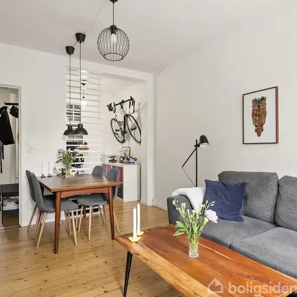 Grå sofa med puder står i stue med lyst trægulv. Ved siden af er der et sofabord med blomster og stearinlys, mens spisebordet har moderne lamper over sig. Væggen har kunst med dyremotiver. Cykler hænger i gangen.