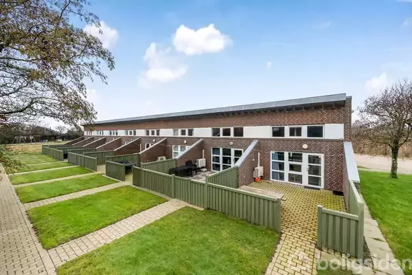 Rækkehuse står i række med grønne græsplæner og gangstier foran, omgivet af grønne hegn. Himlen er klar med få skyer, og bortset fra "Boligsiden" er der ingen tekst.