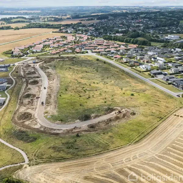 Byområde under opførelse ligger nær kysten og rullende marker. Området omfatter både boliger og tomme byggegrunde. I baggrunden ses en bugt med blå himmel og spredte skyer.
