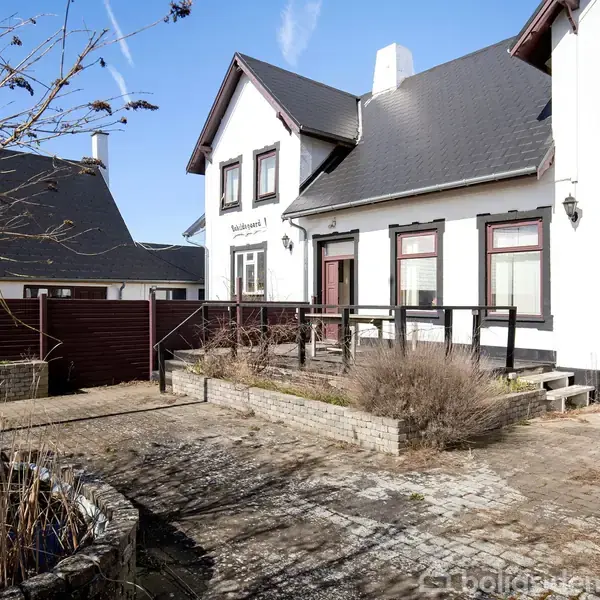 Hvidt hus med sort tag står stille i en gårdhave med flisebelagt område og tørre planter. Sollys skinner fra en blå himmel. Tekst på huset: "Malthuset" og "Anno".