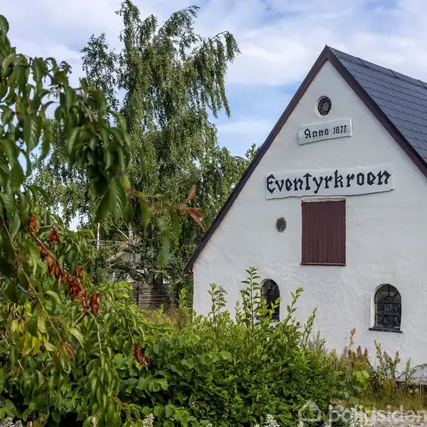 En stor, hvid landbygning står stille med sorte tage. Den er omgivet af flere bygninger og en grusbelagt gårdsplads. Tekst på bygningen: "Andkærgaard".