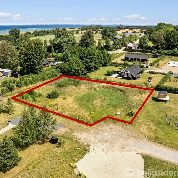 Grunden er omgivet af en rød markering, med græs og buske, beliggende mellem sommerhuse. I baggrunden ses en skov og i horisonten havet under en blå himmel.