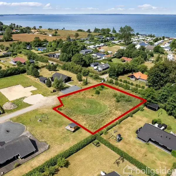 Tomt markeret med rød linje, omgivet af græsmark og huse, beliggende nær en kystlinie. Området består af spredte boliger og landområder med træer og buske i baggrunden.