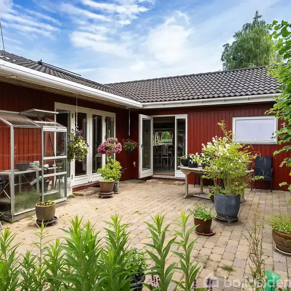Rødmalet hus med åbne dobbeltdøre til en gårdhave. Gårdhaven har flisebelægning, planter i krukker og et lille drivhus. Blå himmel og grønne træer omkranser scenen.