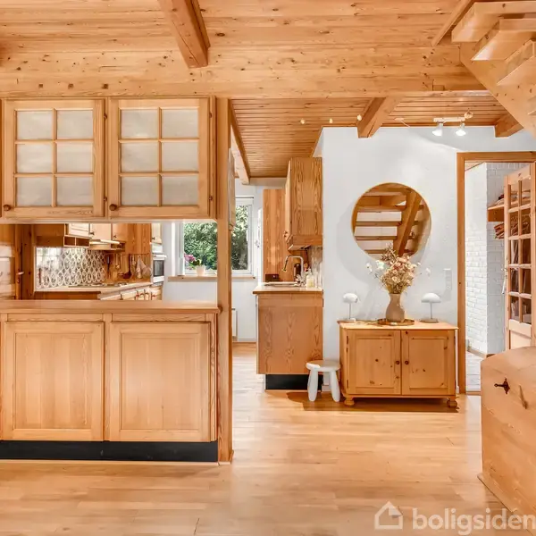 Køkkenet har træskabe og en åben hylde, mens trappen fører ovenpå. Et rundt spejl hænger ved siden af et lille skab med dekorative blomster. Hvidmalede vægge omkranser rummet.