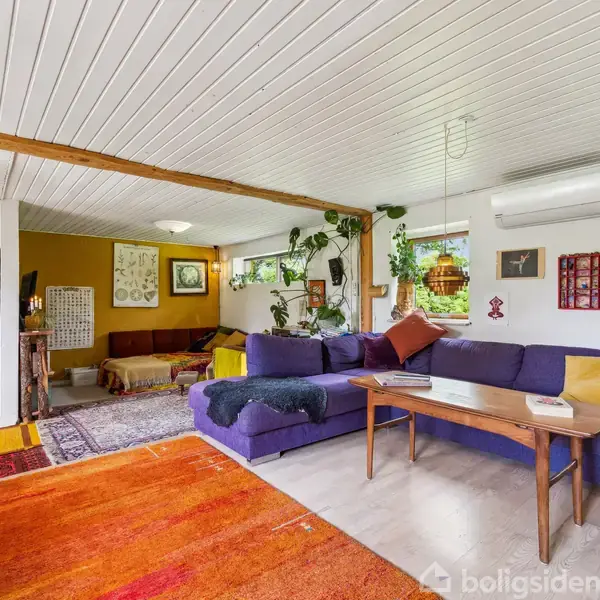 En stor lilla sofa står i midten af en stue, fyldt med farverige puder. Et træbord står foran sofaen. Rummet er lyst med mange planter og en hvid loftbeklædning.