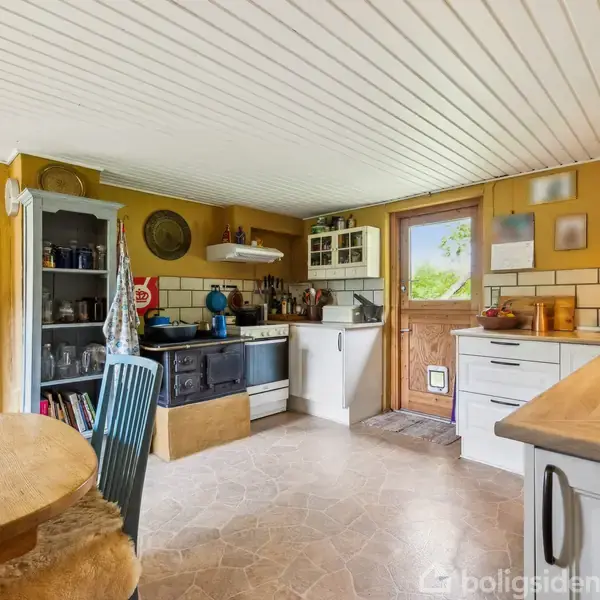 Et køkken med træbord og stole, åbne hylder med glas og krukker, en gammel ovn, hvide skabe, og en dør til haven. Orangerummet ses gennem døråbningen.