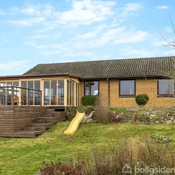 En gul murstensvilla med glaseret veranda forbundet med trappe og gul rutsjebane. Omkring huset er der græsplæne og få buske. Himlen er klar og blå.