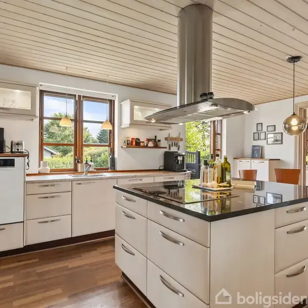 Et moderne køkken med hvidt interiør og en køkkenø med sort bordplade, der har en integreret emhætte. Køkkenet er lyst med træloft og udsigt til en grøn have gennem vinduet.