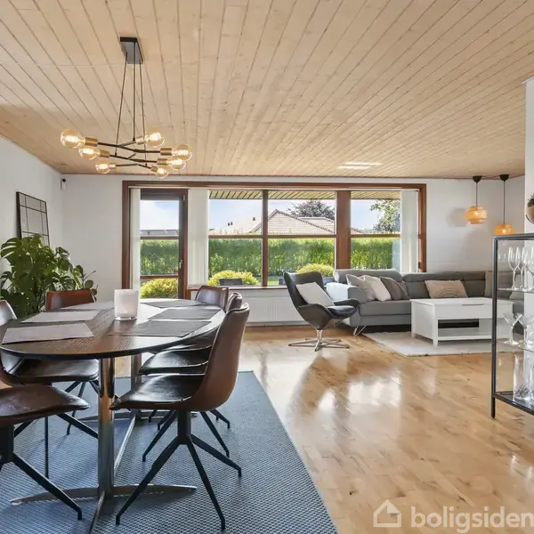 Et spisebord med seks stole står på et tæppe. Store vinduer lukker lys ind i stuen, som har en lysegrå sofa, glasvitrine med glas, træloft og parketgulv.