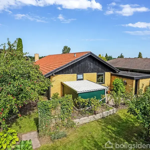 Hus med gult murværk og sort trædetaljer har en overdækket terrasse. Det står i en frodig have med frugttræer. Himlen er blå med få skyer.