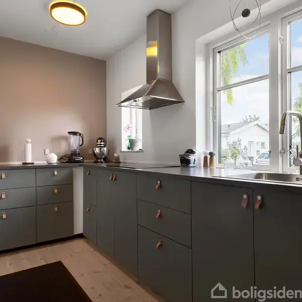 Rødt murstenshus med rødt tegltag og garage ved siden af. Græsplæne og planter omgiver bygningen. Området er en villakvarter med flere huse og marker i baggrunden.