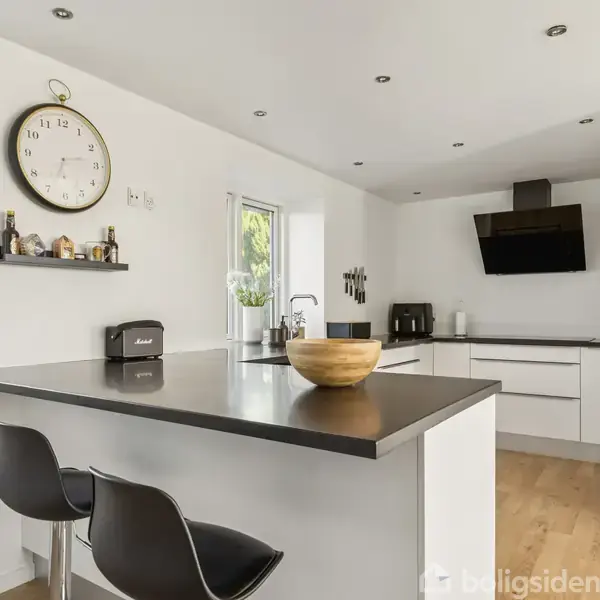 Et moderne køkken med en sort bordplade og to barstole. Hvidt skab, vægur og hylder med krydderier i baggrunden, store vinduer giver naturligt lysindfald.