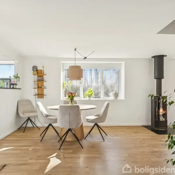 Et rundt bord med fire stole står i en lys stue med trægulv. En brændeovn er placeret til højre, og en vindueskarm har grønne planter.