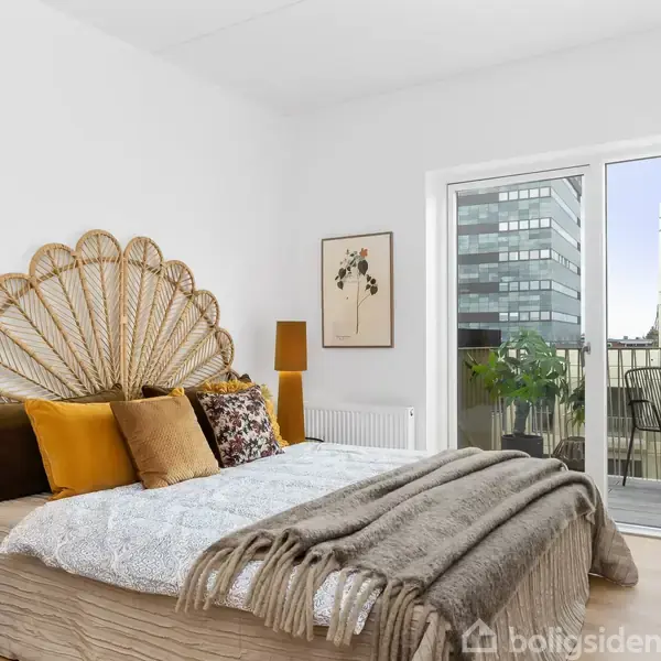 Soveværelse med seng og farverige puder, placeret ved siden af en glasdør, der fører til altan med udsigt over bybygninger. En plante og en stol står på altanen.