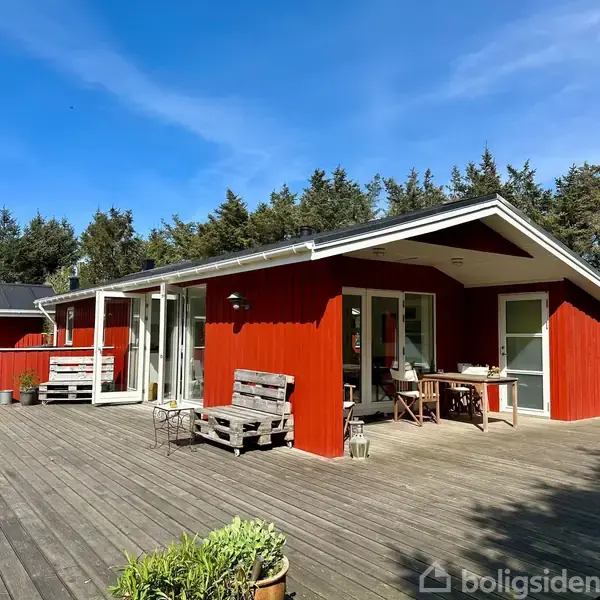 Rødt sommerhus med store vinduer står på en træterrasse. Der er havemøbler foran huset. Omgivet af grønne træer og græsplæne under en klar blå himmel.