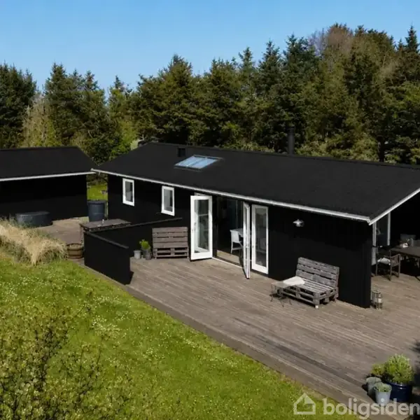 Sort træhus står stille på en træterrasse, omgivet af en frodig have med grønne træer. Huset har store vinduer og en tilhørende mindre bygning.