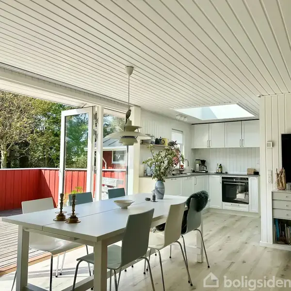 Spisebord med stole står centralt i et lyst køkken-alrum. Rummet har åbne glasdøre til en terrasse med rødmalet hegn. Køkkenet er moderne med hvide skabe og ovenlys.