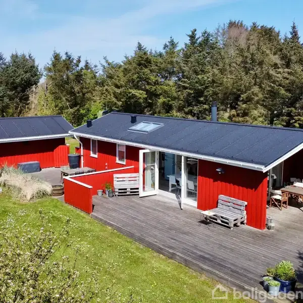 Rødt træhus står på en stor træterrasse, omgivet af en frodig have med græsplæne og træer. Vinduer og døre har hvide karme; sollys oplyser området.