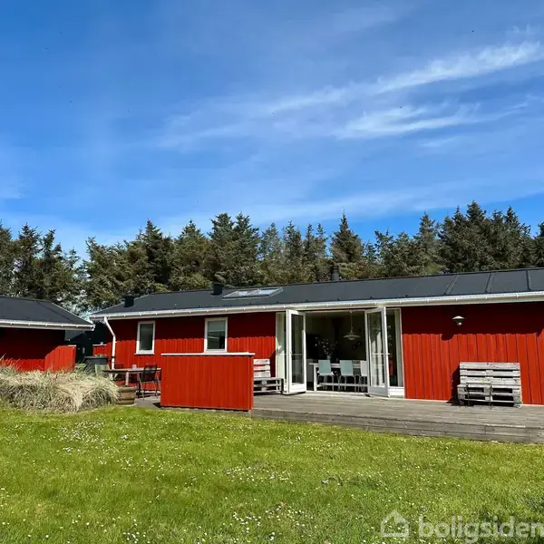 Rødt sommerhus med sorte tage står stille i en grøn have omgivet af tæt skov. Terrassen foran huset har hvide havemøbler under en klar, blå himmel.