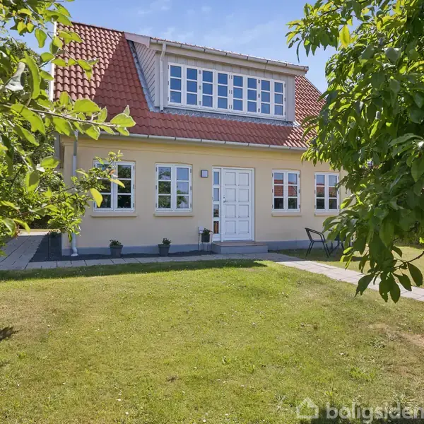 Et to-etagers hus med rødt tegltag har en hvid dør og seks vinduer. Det står i en grøn have med træer og buske under en klar himmel.