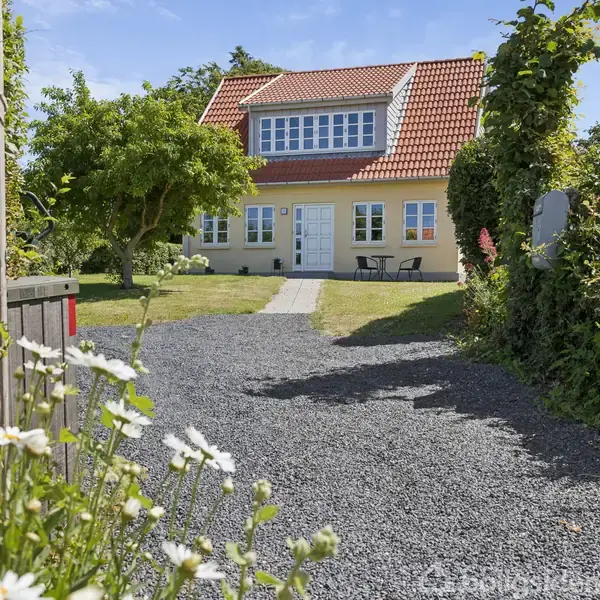 Et gulmalet hus med rødt tegltag står i en frodig have med grusgang og blomster. Et træ skaber skygge nær huset, der har hvide vinduer og døre.