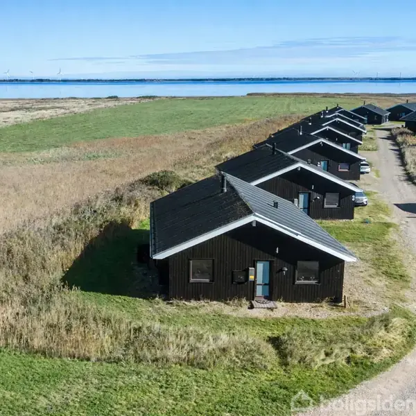 Række af sorte træhuse ligger på en grusvej ved en strandeng. Husene står side om side med udsigt til en blå himmel og vand i baggrunden.
